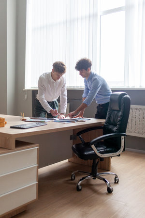 young male boss in business clothes sitting in office at table and talking with colleague at work, entrepreneur and his partner smiling and discussing report and planの写真素材