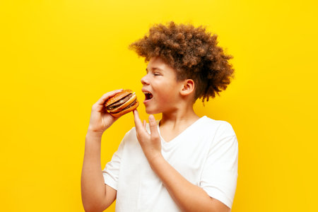 cheerful curly african american boy eating burger on yellow isolated background, hungry teenager...