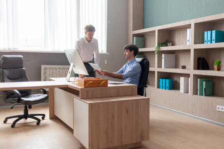 young male boss in business clothes sitting in office at table and talking with colleague at work, entrepreneur and his partner smiling and discussing report and planの写真素材