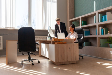 African American woman and European young man in business suit using computer and talking in office,...