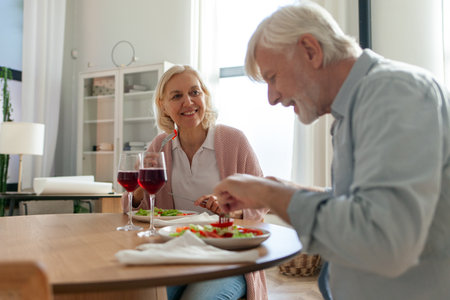 elderly couple is having lunch at home at the table and talking, gray-haired husband and wife are celebrating and drinking red wine and eating saladの写真素材