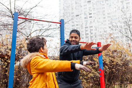 African American man and his son exercise and do exercises outdoors on playground, teenager stretches with his father on exercise equipmentの写真素材