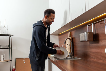 African American man washes dishes alone in the kitchen, guy cleans the house and does houseworkの写真素材