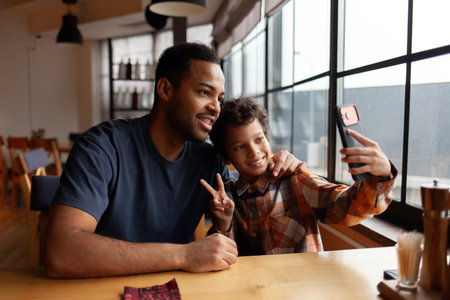 African American man and his son take selfie on smartphone in cafe at table, teenager and his father sit in restaurant and take photo together on mobile phoneの写真素材