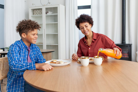 African American woman eating chocolate butter banana sandwiches and juice with her son at home at the table, teenager having lunch with his mother and laughingの写真素材