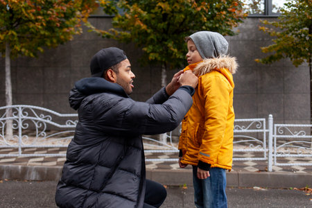 African American man taking care and dressing his son in warm jacket and hat outdoors, father buttoning his childs shirt and checking warm clothes in autumn in the cityの写真素材