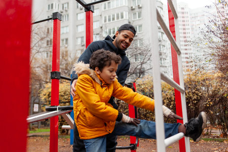 African American man and his son exercise and do exercises outdoors on playground, teenager stretches with his father on exercise equipmentの写真素材