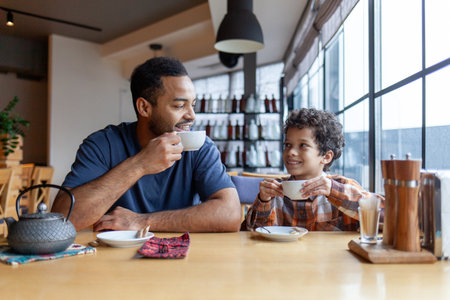 African American man and his son are having lunch in cafe at table, teenager and his father are sitting in restaurant and eating and drinking coffeeの写真素材