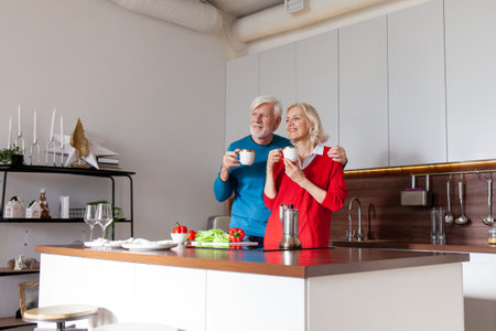 elderly couple of seniors drink coffee in the kitchen and smile, old woman and man hold cups of tea...