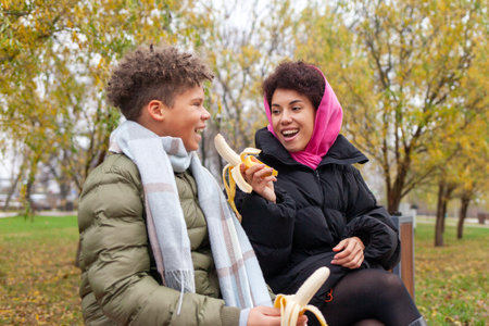 African American woman and son eating banana in park in autumn, teenager sitting with mother outdoors in warm clothes and eating lunchの写真素材