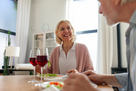 elderly couple is having lunch at home at the table and talking, gray-haired husband and wife are celebrating and drinking red wine and eating saladの写真素材