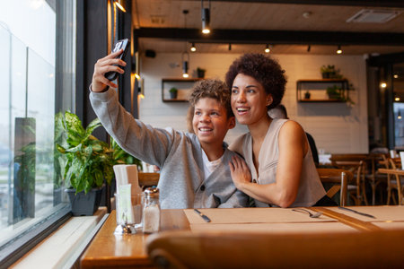 African-American woman and her son are sitting in cafe and taking selfies on smartphone, teenager and his mother are taking photos on phone in restaurantの写真素材