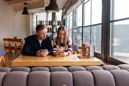 young couple is sitting at table in cafe and using smartphone, girl and guy are drinking tea in...