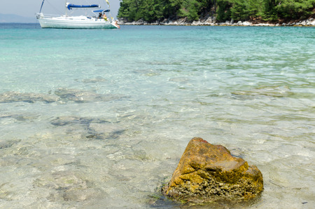 Seascape view with a rock in foreground and a yacht in a bay with tourquoise waterの写真素材