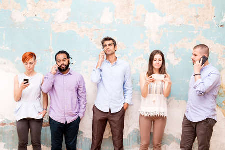 Group of young people standing in front of a grunge wall with a mobile phone handsの写真素材