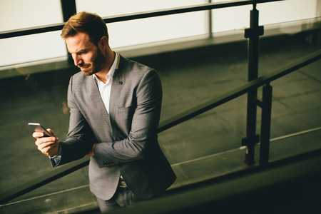 Young successful businessman wearing grey suit and holding his smartphone while standing near modern office or skyscrapersの写真素材