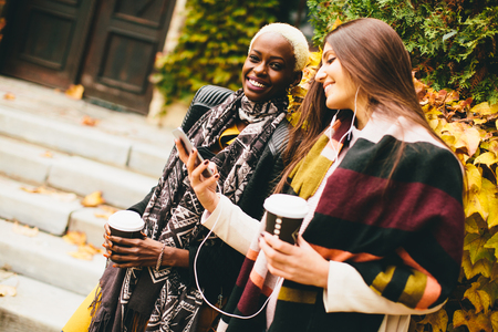African american and caucasian woman posing outside with mobile phone and a cup of coffee to go in autumnの写真素材