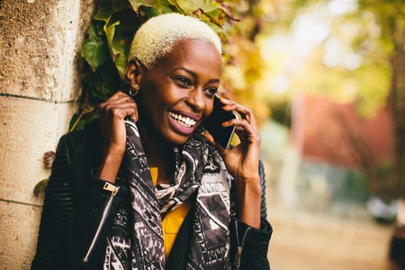 Modern young african american woman holding mobile hone and smiling at autumn outdoorの写真素材