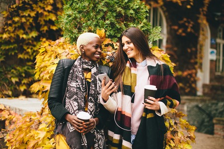 African american and caucasian woman posing outside with mobile phone and a cup of coffee to go in autumnの写真素材