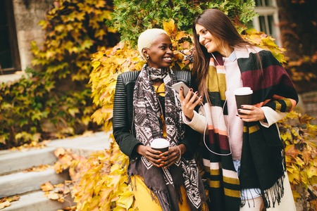 African american and caucasian woman posing outside with mobile phone and a cup of coffee to go in autumnの写真素材
