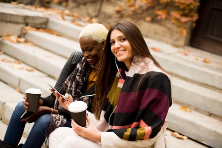 African american and caucasian woman posing outside with mobile phone and a cup of coffee to go in autumnの写真素材