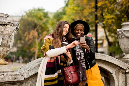 African american and caucasian woman posing outside with mobile phone and a cup of coffee to go in autumnの写真素材