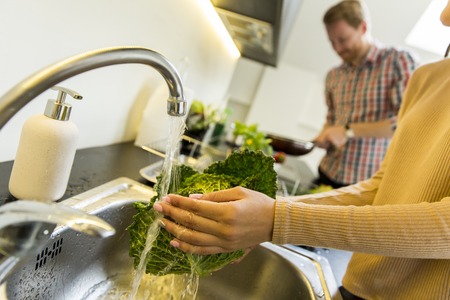 Young woman washes kale on the tap while a young man in the background cookingの写真素材