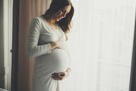 Pretty young pregnant woman standing by the window in roomの写真素材