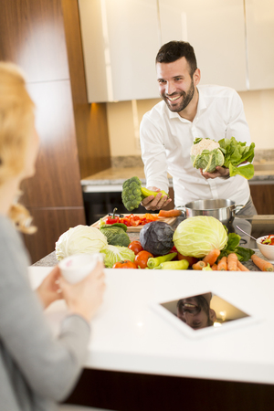 Young woman sitting in the kitchen and drinking coffee while a young man preparing vegetablesの写真素材