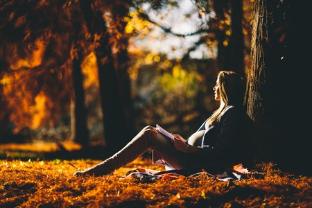 Beautiful pregnant woman sitting under tree , with pleasure expecting  baby and having happy pregnancy timeの写真素材
