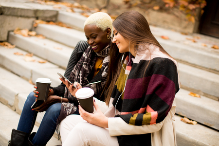 Young multiracial friends sitting in autumn park, talking and carry coffee to go in the handsの写真素材