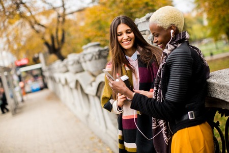 Two young pretty multiracial girlfriends drinking coffee and using mobile phone at autumn dayの写真素材