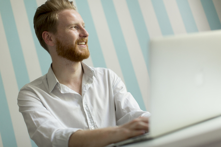 Portrait of young redhair man working on laptop in room by the blue striped wallの写真素材