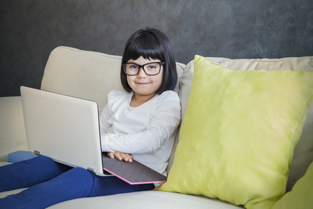Beautiful little girl with glasses using her laptop while sitting on the couch at home
