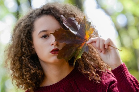 Portrait of curly hair teen girl holding maple leaf in autumn forestの写真素材