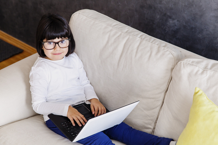 Cute little girl in casual clothes using a laptop while sitting on sofa at homeの写真素材