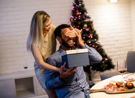 Portrait of happy woman closing male eyes by her hands while holding giftbox in front of himの写真素材
