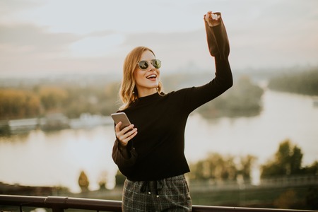Portrait of young woman holding mobile phone while standing on walkway by the riverの写真素材