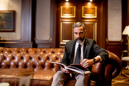 Handsome middle-aged businessman reading book on sofa