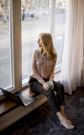 Pretty young woman using laptop and sitting by windowの写真素材