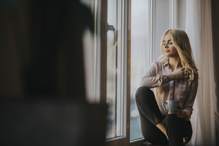 Blonde young woman relaxing and  drinking coffee by windowの写真素材