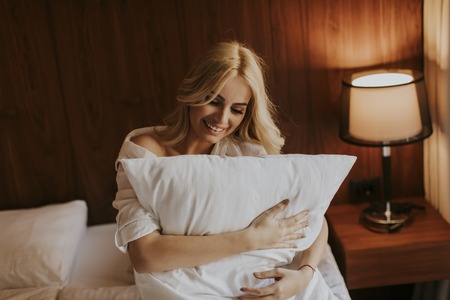 Portrait of happy blonde woman hugging her pillow at home in the bedroomの写真素材