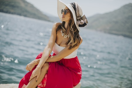 Pretty young woman with hat and sunglasses sitting by the seaの写真素材