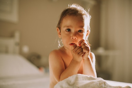 Sweet curly little girl with a towel over her wet body siting in a bedroom after shower or bathの写真素材