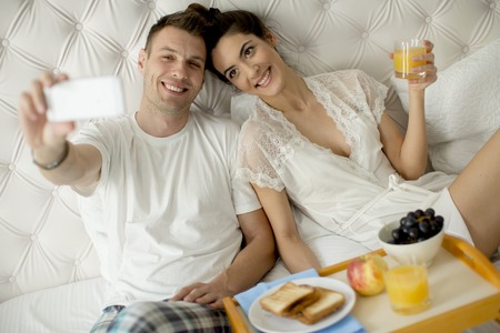 Young couple having a breakfast in the bed and taking selfie with mobile phoneの写真素材