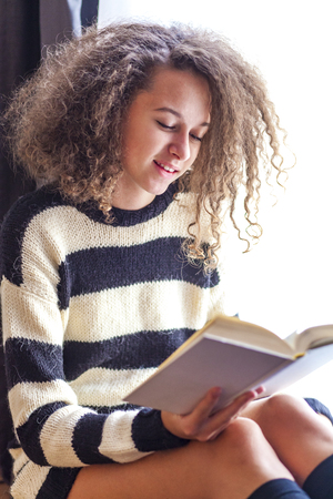 Curly hair teen girl reading book by windowの写真素材