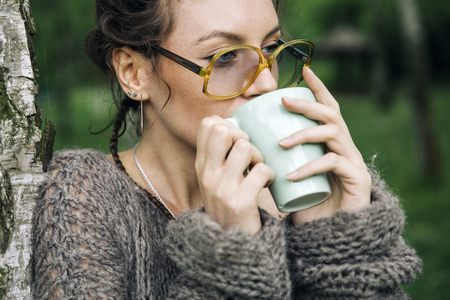 Portrait of young woman in a sweater and with glasses drinking coffee in the parkの写真素材