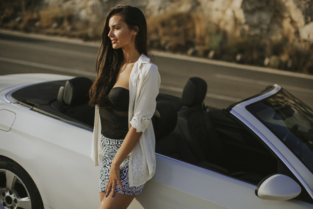 Pretty young woman standing by the white cabriolet car on a sunny day by the seaの写真素材