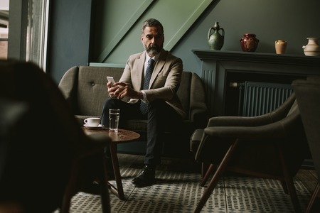 Portrait of handsome senior businessman drinking coffee and using mobile phone in lobbyの写真素材