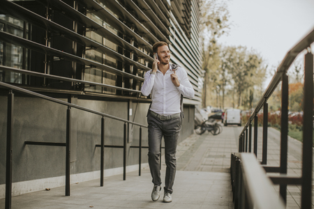 Handsome young businessman walking with mobile phone in front of office buildingの写真素材